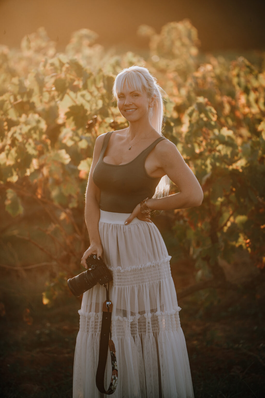 Portrettbilde av Fotograf Lene Fossdal. Hun holder et kamera i ene hånden, den andre i siden. Hun ser i kamera og smiler. Hun står foran en vinåker, og solen er bak henne. Hun smiler og ser i kamera.